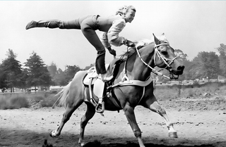 Person riding a horse with one leg extended, set against a natural background.